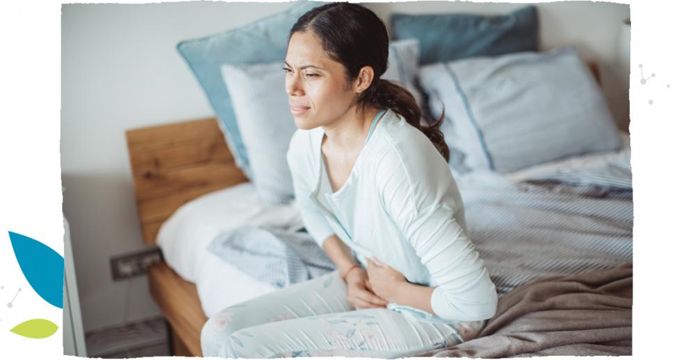 Žena v modrém pyžamu sedí na posteli a trpí bolestmi žaludku
