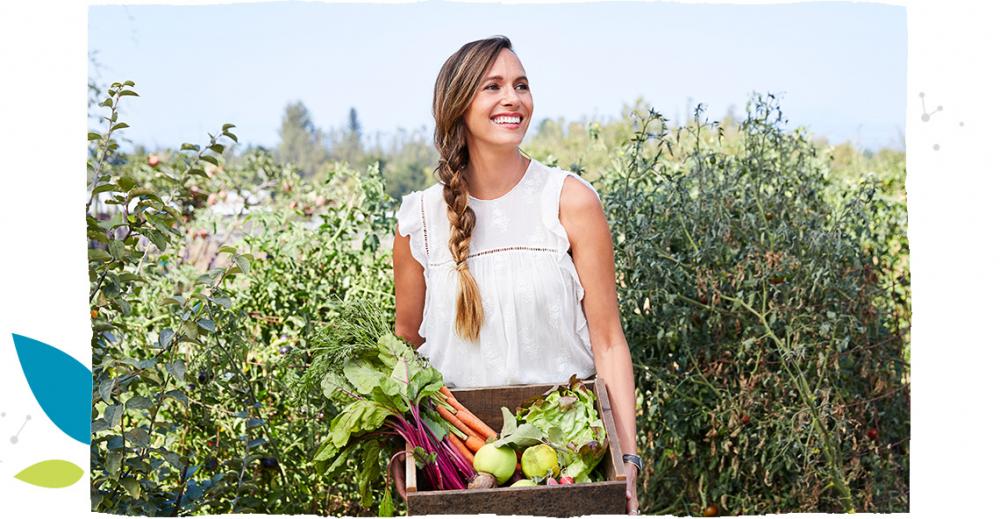 Žena stojí uprostřed pole a drží přepravku plnou zeleniny