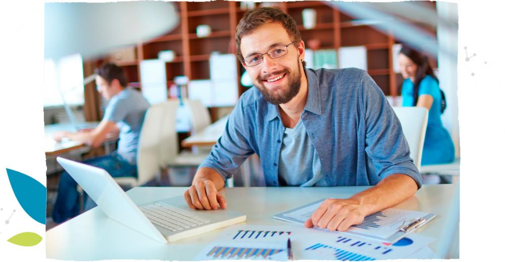 Usměvavý vousatý mladý muž s brýlemi pracuje na svém počítači s papíry ležícími na jeho bílém stole